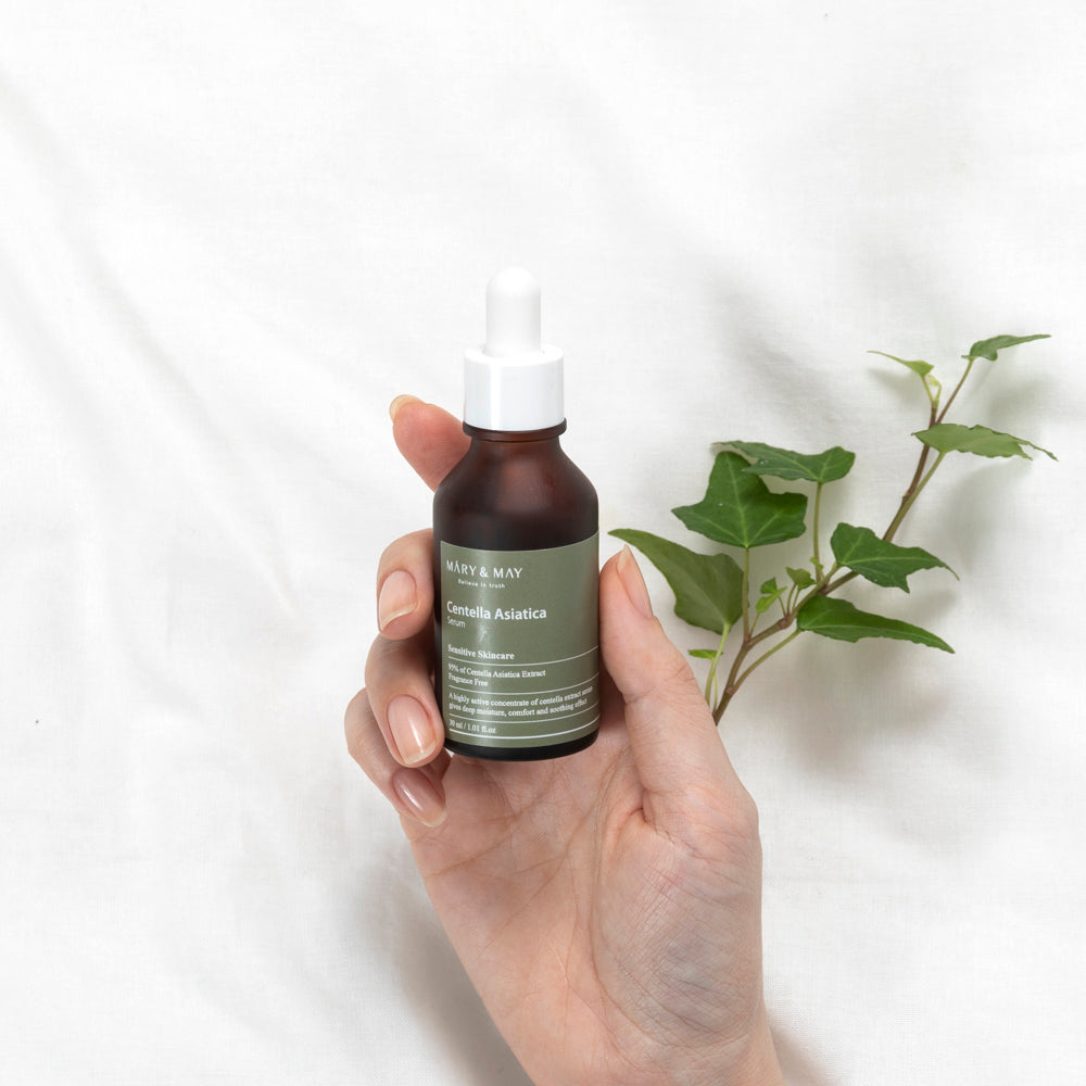 Hand holding a bottle of 'Mary & May' serum with a white background and green leaves.