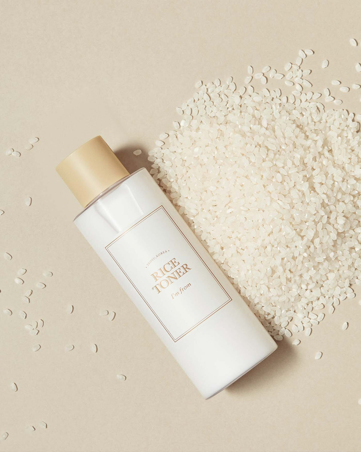 White bottle of rice toner with a beige cap on a beige background with scattered rice grains.